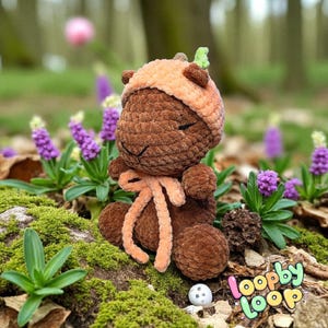 May include: A handmade brown crochet bear wearing an orange hat and scarf, with a small green leaf accent. The bear is sitting among purple flowers and green moss. The LoopbyLoop logo is visible in the bottom right corner.