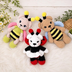May include: Three handmade crochet bee-themed soft toys. One is white with red accents, another is tan, and the third is pink. Each has a bear-like face, wings, and a striped body. The toys are on a white, fluffy surface.