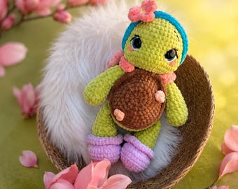 Puede incluir: Una muñeca de tortuga de ganchillo con cuerpo verde, caparazón marrón y zapatos morados. La tortuga lleva una diadema azul con una flor rosa y una bufanda rosa. Está sentada en una cesta tejida sobre una cama de piel blanca, rodeada de flores de magnolia rosas.