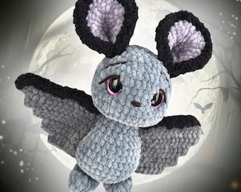 May include: A gray crocheted bat with black ears and wings. The bat has pink eyes and a cute expression. The bat is flying in front of a full moon.
