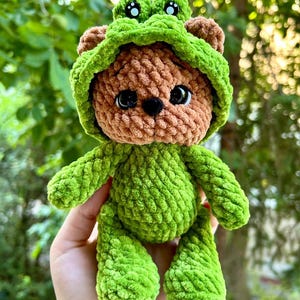 May include: A handmade crochet teddy bear wearing a green frog costume. The bear is brown with black eyes and a black nose. The frog costume includes a hood with frog eyes and a green body. The bear is being held in a hand.