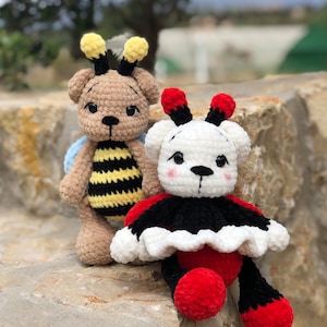 May include: Two crocheted teddy bears dressed as a bee and a ladybug. The bee bear is brown with yellow and black stripes and blue wings. The ladybug bear is white with black spots and a red skirt.