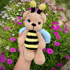 May include: A crocheted bee-themed teddy bear with brown fur, yellow and black striped body, and blue wings. The bear is held in a hand against a background of pink and white flowers.