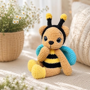 Op de afbeelding: Een handgemaakte gehaakte teddybeer met een bijenthema. De beer heeft een bruin lichaam met zwarte en gele strepen, blauwe vleugels en gele voeten. Hij heeft een zwarte haarband met gele antennes en grote blauwe ogen. De beer zit op een lichtgekleurd oppervlak.