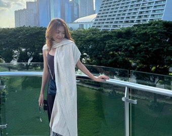 Puede incluir: Una mujer con una larga bufanda de color crema sobre un vestido negro sin mangas. Está de pie en un balcón con una barandilla de vidrio y una vista del horizonte de la ciudad.