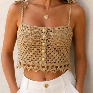 Peut inclure: Un haut court en crochet beige avec des boutons dorés et des bretelles nouées. Le haut a une encolure carrée et un ourlet festonné. Le modèle porte un pantalon blanc et des bijoux dorés.