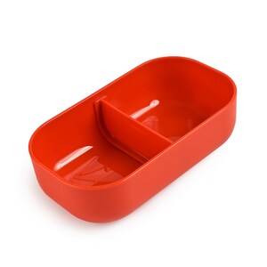 May include: A vibrant red food container, rectangular with rounded edges, and a central divider. The container is made of a smooth, solid material, designed to hold two separate food items. The container is empty and sits against a white backdrop.