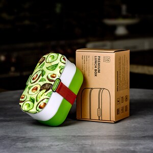 May include: Green and white lunch box with a colourful avocado print. The lunch box has a red and green strap and is made of durable plastic. The box is sitting on a grey surface next to a brown box with the text "PREMIUM LUNCH BOX" printed on it.