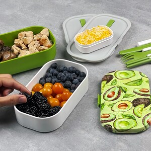 May include: A set of four green and white lunch containers with a variety of food inside, including mushrooms, chicken, blueberries, tomatoes, and blackberries. The containers are arranged on a grey surface with a set of green utensils and a brown strap with a metal buckle.
