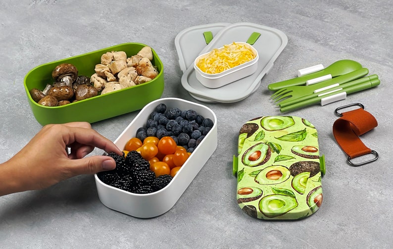 May include: A set of four green and white lunch containers with a variety of food inside, including mushrooms, chicken, blueberries, tomatoes, and blackberries. The containers are arranged on a grey surface with a set of green utensils and a brown strap with a metal buckle.