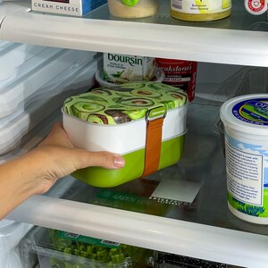 May include: A green and white lunch box with a brown strap and an avocado print. The lunch box is labelled "Spillproof" and "Green Leaf" and is sitting in a refrigerator.