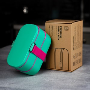 May include: A teal and pink lunch box with a strap and the word "Hämtmat" on the side. The lunch box is sitting on a gray surface next to its brown cardboard box. The box has the text "PREMIUM LUNCH BOX" printed on it.