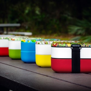 May include: A row of colourful lunch boxes with a variety of patterns. The lunch boxes are stacked on top of each other and have a black strap.