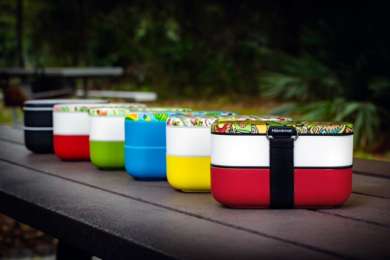 May include: A row of colourful lunch boxes with a variety of patterns. The lunch boxes are stacked on top of each other and have a black strap.