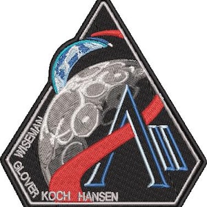 May include: Embroidered patch with a black triangular shape, depicting a planet and a large letter "A". The patch includes the names "WISEMAN", "GLOVER", "KOCH", and "HANSEN". The design incorporates red, white, and blue colours.