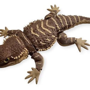 Peut inclure: Figurine de lézard marron et beige détaillée, avec un corps texturé et des détails épineux. Le reptile a une tête détaillée avec des yeux jaunes et est posé sur un fond blanc. Les pattes du lézard sont écartées et sa queue est étendue.