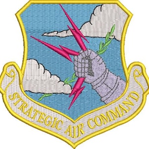 May include: Embroidered patch featuring a shield design with a light blue background and a gold border. The patch includes a stylized armored fist holding pink lightning bolts, with the text "STRATEGIC AIR COMMAND" in gold.