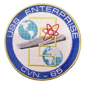 May include: Circular embroidered patch with the text "USS ENTERPRISE" in blue, and "CVN-65" below. The patch has a white centre with a yellow outline, and a blue border. The design includes two globes, a grey aircraft, and a red atom symbol.