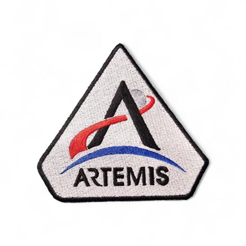 Pu&ograve; includere: Toppa ricamata con il logo del programma Artemis. Il design include una 'A' nera con un tratto rosso e bianco, un arco blu e la parola 'ARTEMIS' in bianco. La toppa &egrave; un triangolo bianco con un bordo nero.