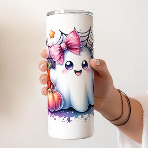 Pu&ograve; includere: Un bicchiere in acciaio inossidabile bianco con un fantasma dei cartoni animati che indossa un fiocco rosa e tiene una zucca. Il fantasma ha occhi grandi e un'espressione felice. Lo sfondo &egrave; un design ad acquerello viola e bianco con una ragnatela.