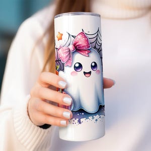 Pu&ograve; includere: Un bicchiere in acciaio inossidabile bianco con un fantasma dei cartoni animati che indossa un fiocco rosa e circondato da ragnatele. Il fantasma ha occhi grandi e un'espressione felice.