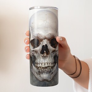 May include: A tall, cylindrical tumbler featuring a detailed, realistic illustration of a human skull. The skull is rendered in shades of white, gray, and brown, with visible teeth and eye sockets. The design is printed on the tumbler's surface.