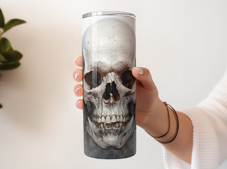 May include: A tall, cylindrical tumbler featuring a detailed, realistic illustration of a human skull. The skull is rendered in shades of white, gray, and brown, with visible teeth and eye sockets. The design is printed on the tumbler's surface.
