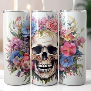 May include: White stainless steel tumbler featuring a detailed skull design adorned with colorful flowers. The floral arrangement includes pink roses, blue and purple peonies, and green foliage. The tumbler has a clear lid and a straw.