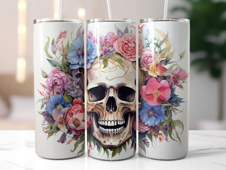 May include: White stainless steel tumbler featuring a detailed skull design adorned with colorful flowers. The floral arrangement includes pink roses, blue and purple peonies, and green foliage. The tumbler has a clear lid and a straw.