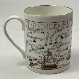 May include: A white ceramic mug with a gold-trimmed rim featuring a colorful map illustration titled THE NORMAN COAST. The design includes names of locations like Cilgerran, Haverfordwest, and Tenby, alongside small castle icons and historical figures.