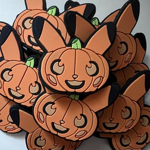May include: A collection of orange and brown ceramic pumpkin-shaped ornaments with a cartoon-like face. The ornaments have a glossy finish and are decorated with black eyes, a black mouth, and a black nose.