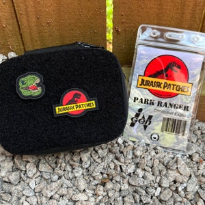 May include: A black pouch with two Jurassic Park patches, one featuring a dinosaur and the other with the text "Jurassic Patches". A clear plastic badge holder with the text "Park Ranger Dinosaur Expert" and a Jurassic Park logo.