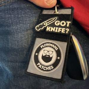 以下が含まれることがあります： 「Plowmans Patches」という文字と、眼鏡をかけたひげを生やした男性の漫画の絵が描かれた白黒のパッチ。パッチは、銀色のキーリングが付いた黒と灰色のキーホルダーに取り付けられています。キーホルダーには、ナイフの画像と「Got Knife?」という文字が書かれた白黒のパッチも付いています。