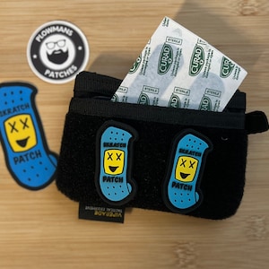 以下が含まれることがあります： 黒いジッパー付きポーチに、青い顔文字のばんそうこうが2つ付いています。ポーチが開いていて、中から滅菌されたばんそうこうの包みが覗いています。木製の表面には、「Plowmans Patches」と書かれた丸いステッカーが見えます。