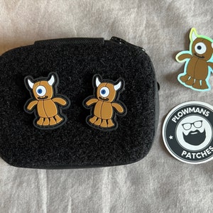 Könnte beinhalten: Zwei braune Monster-Patches mit jeweils einem blauen Auge, befestigt an einem schwarzen Molle-Panel. Ein holografischer Aufkleber eines braunen Monsters mit einem blauen Auge und dem Text "Plowmans Patches" auf weißem Hintergrund.