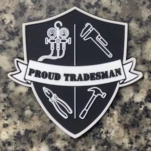 Op de afbeelding: Een zwart-wit schildvormig embleem met de tekst "PROUD TRADESMAN" op een banner. Het schild toont lijntekeningen van gereedschappen: een meter, een tang, een hamer en een mechanisch apparaat.