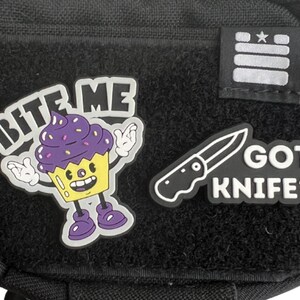 May include: Black pouch with three white patches. The patches have the text "Bite Me" with a cartoon cupcake, "Got Knife?" with a knife, and three white stripes with a star.