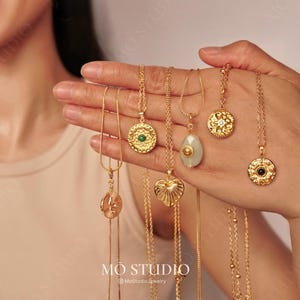 Peut inclure: Un ensemble de colliers en or avec différents pendentifs, dont un cœur, un cercle avec une pierre verte, un cercle avec une pierre noire, un cercle avec une pierre blanche et un pendentif en forme de larme avec une pierre vert clair. Les colliers sont présentés sur une main.