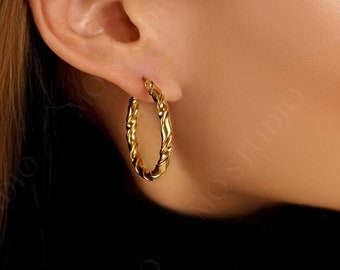May include: A close-up of a woman's ear wearing a pair of gold hoop earrings. The earrings are thick and have a twisted design.