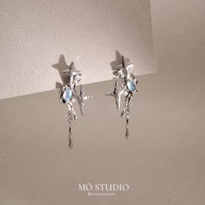 Peut inclure: Boucles d'oreilles argentées au design céleste. Chaque boucle d'oreille présente une étoile, un pendant en forme de goutte et une pierre irisée. Les boucles d'oreilles sont de MO STUDIO.