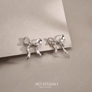 May include: A pair of silver bow earrings. Each earring has a detailed bow design with flowing ribbon-like elements. The earrings are displayed on a neutral surface, with the brand name "MO STUDIO" visible at the bottom.