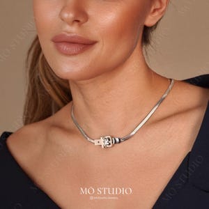 Puede incluir: Un collar de cadena de plata con cierre de hebilla de plata. El collar está puesto por una modelo que lleva una camisa negra.