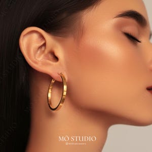 May include: Gold hoop earrings with a textured surface. The earrings are worn by a person with their eyes closed.