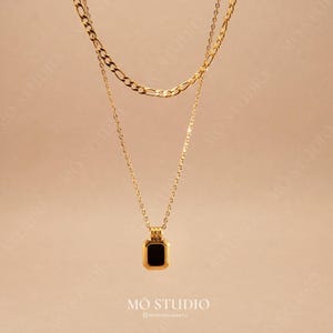 Pode incluir: Um colar de corrente de ouro com um pingente quadrado com uma pedra preta. O colar é sobreposto com uma corrente de ouro mais grossa.