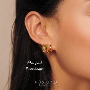 Può includere: Primo piano di un orecchio con tre orecchini a cerchio dorati. Gli orecchini sono impilati uno sopra l'altro, con il cerchio più grande in basso e il più piccolo in alto. Il testo "One post, three hoops" è scritto sotto gli orecchini.