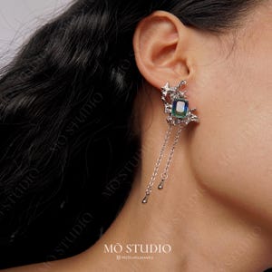 May include: Silver-toned earring with a rectangular, iridescent gemstone and dangling chain accents. The earring features a star-shaped design and the text "MŌ STUDIO" at the bottom.
