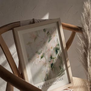 May include: A framed art print featuring a soft, impressionistic floral design in pastel pinks and greens. The print is titled "R&ecirc;ve de Pivoines" and is displayed on a wooden chair with a neutral-toned fabric. A dried ornamental grass adds a natural element.