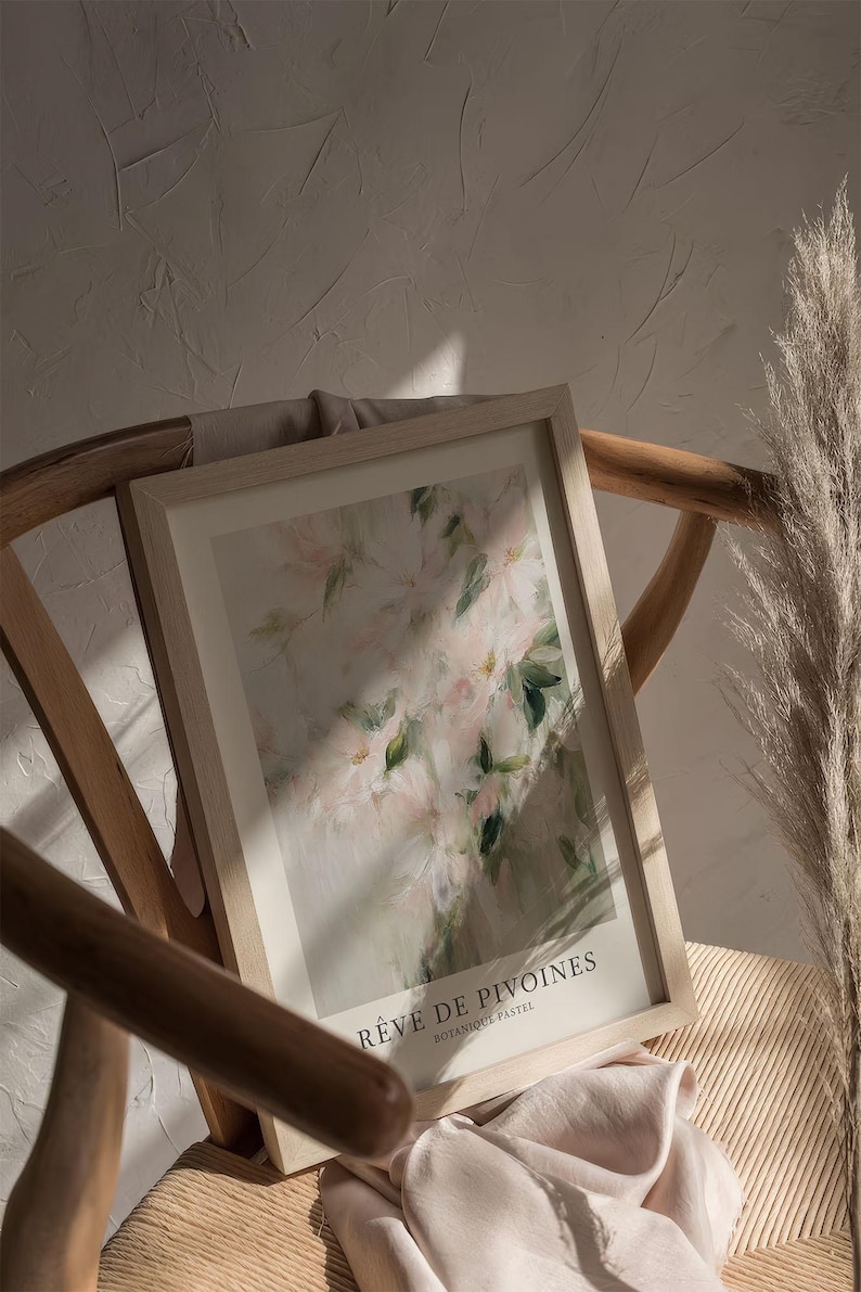 May include: A framed art print featuring a soft, impressionistic floral design in pastel pinks and greens. The print is titled "R&ecirc;ve de Pivoines" and is displayed on a wooden chair with a neutral-toned fabric. A dried ornamental grass adds a natural element.