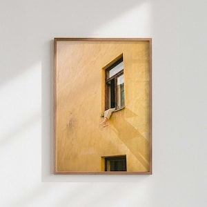 Op de afbeelding: Een ingelijste foto van een geel gebouw met een raam. De benen van een persoon zijn te zien die uit het open raam hangen. De raamkozijn is bruin. De foto is aan een witte muur bevestigd.