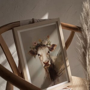 May include: A framed painting of a white cow wearing a flower crown. The cow is looking directly at the viewer with a gentle expression. The background is a light brown colour.